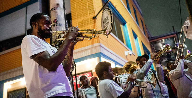 A close-up of a brass band musician playing a trumpet on a vibrant New Orleans street, with historic French Quarter architecture in the background.