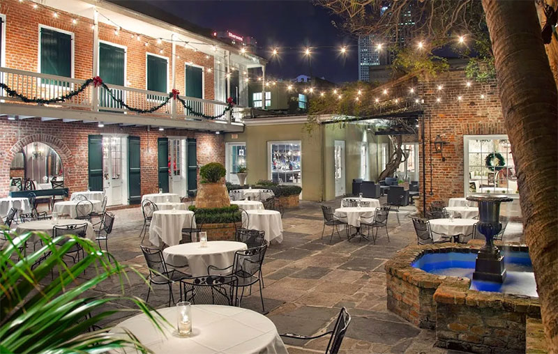 A view of the lush, historic outdoor courtyard at Broussard’s Restaurant in the French Quarter, featuring large tropical plants, white-clothed dining tables, and classic architecture under the open sky.
