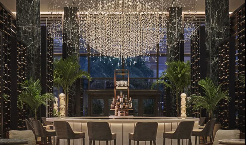 A sophisticated interior view of the Chandelier Bar at the Four Seasons Hotel New Orleans, featuring a massive, shimmering crystal chandelier centerpiece over a modern circular bar with plush seating.