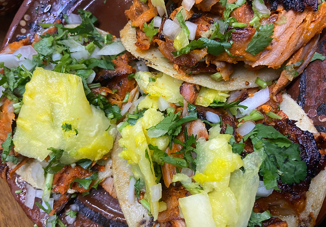 Close-up of Al Pastor tacos topped with pineapple slices, chopped onions, and cilantro on corn tortillas.