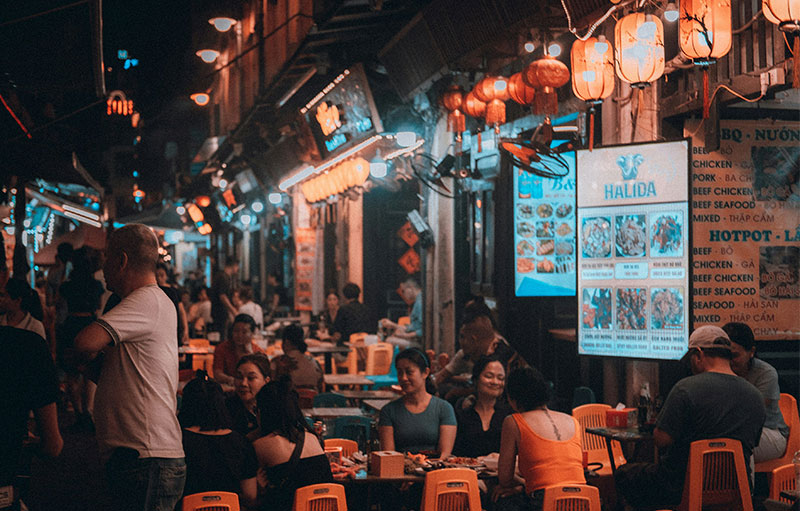 A busy night market scene in Hanoi, Vietnam, with people sitting on low plastic stools at outdoor food stalls under glowing red lanterns and warm street lights