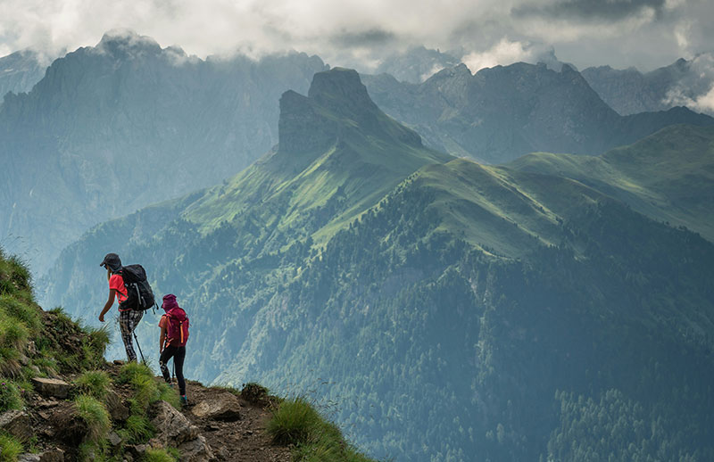 Two hikers with backpacks stand on a grassy mountain ridge in the Dolomites, Italy, looking out over jagged, dramatic rock peaks under a soft, hazy sky.