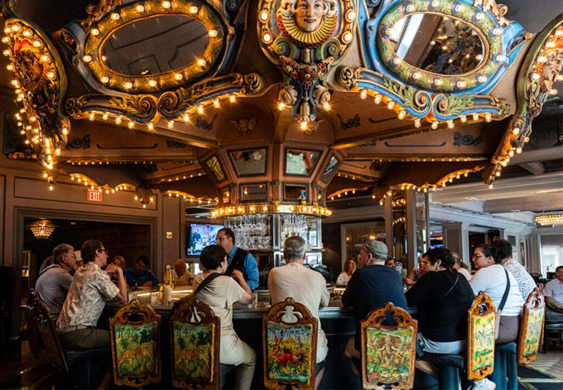 A wide interior shot of the historic Carousel Bar at Hotel Monteleone in New Orleans, featuring the ornate, rotating circus-themed bar with glowing lights and patrons seated on colorful stools.