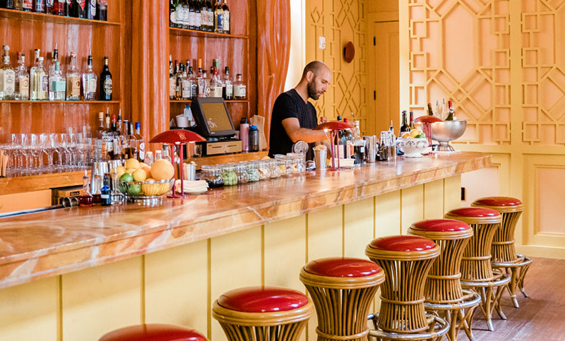 A warm and inviting interior view of The Elysian Bar at Hotel Peter & Paul, featuring a long wooden bar with red-cushioned stools, ornate yellow-toned walls, and a large arched mirror.