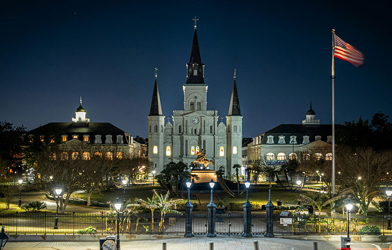 A wide night shot of Jackson Square in New Orleans, featuring the illuminated St. Louis Cathedral with its three spires against a dark sky, framed by the park's greenery and iron fencing.