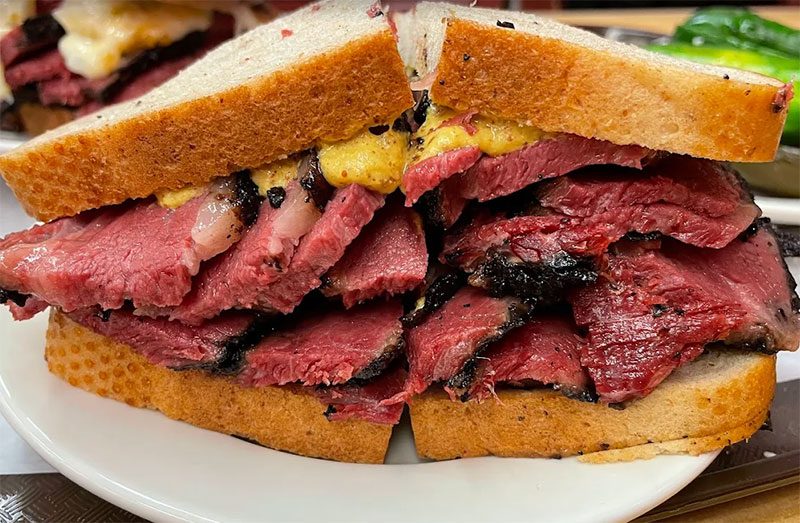 A close-up of a massive Katz’s Delicatessen pastrami sandwich on rye bread, piled high with juicy, hand-carved meat and served with a side of classic deli pickles.