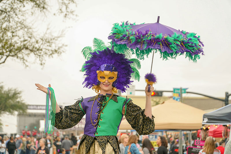 A person dressed in an elaborate, bright purple and green Mardi Gras costume with a feathered headpiece, holding a matching decorated umbrella and posing on a New Orleans street.