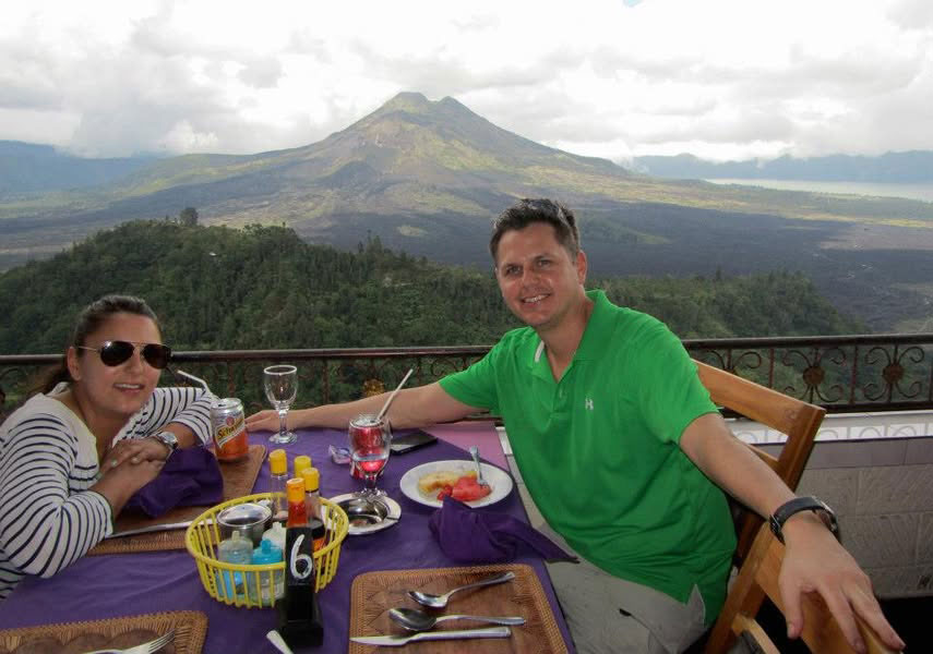 Corey in a green polo shirt and a woman in a black top sit at an outdoor restaurant table with a spread of food, overlooking a vast tropical valley and the distant volcanic peaks of Mount Batur in Kintamani, Bali.
