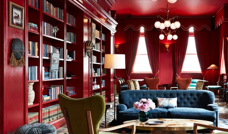 An interior view of the bar at Maison Métier hotel, featuring a luxury design with deep red walls, floor-to-ceiling bookshelves, velvet armchairs, and elegant dim lighting.