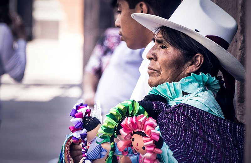 An elderly indigenous Mexican woman wearing a traditional purple and green garment and a white hat, looking off-camera with a serious expression.