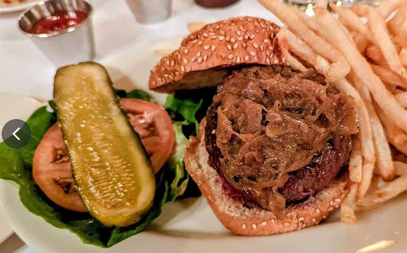 A close-up of the famous Black Label Burger at Minetta Tavern, served on a wooden board with a side of thick-cut pickles and pommes frites.