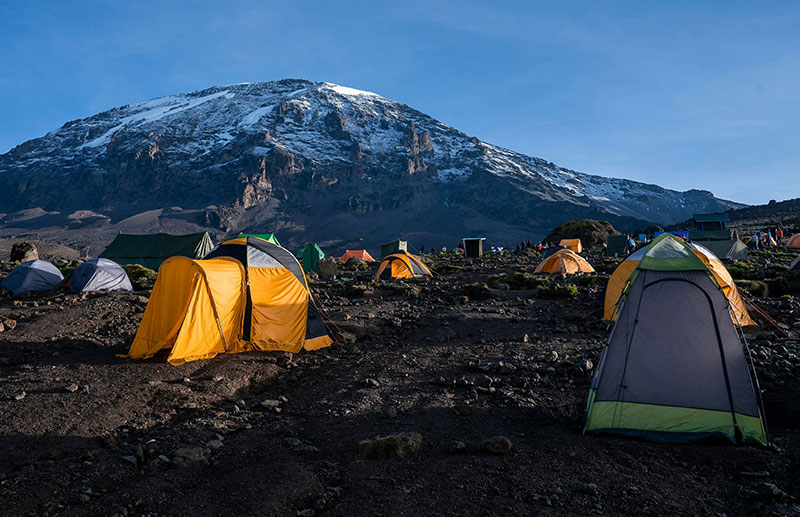 A group of hikers with backpacks trekking up a rocky path toward the snow-capped peak of Mount Kilimanjaro during a golden sunrise.
