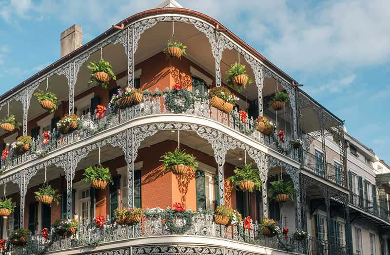 A vibrant two-story historic building in the New Orleans French Quarter with wrap-around wrought-iron balconies, overflowing hanging flower baskets, and classic shuttered windows.