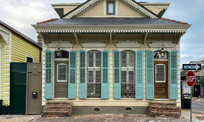 A classic New Orleans "shotgun" style house with a light green facade, decorative cream-colored trim, and traditional turquoise shutters.