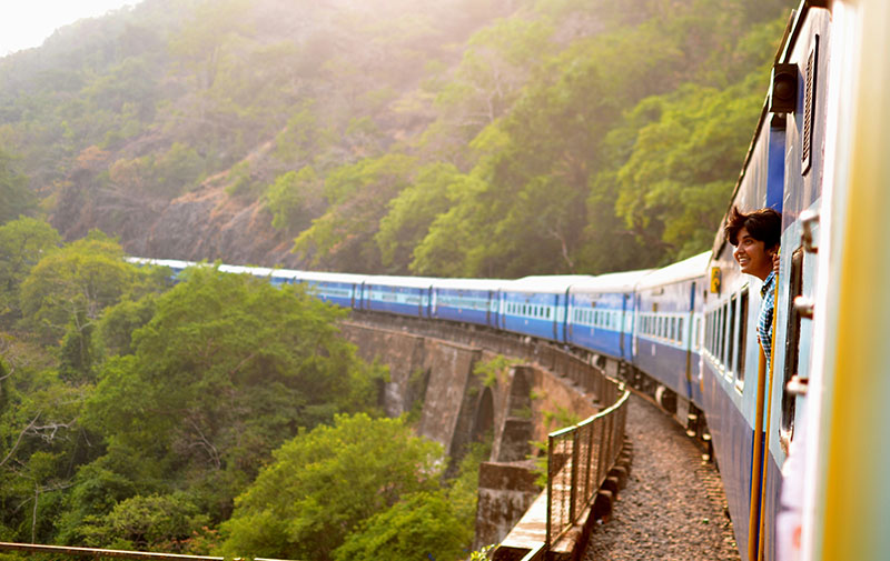 A person looks out from the open door of a blue and white passenger train as it travels along a curved track over a stone arched bridge, surrounded by lush green forested hills in India.