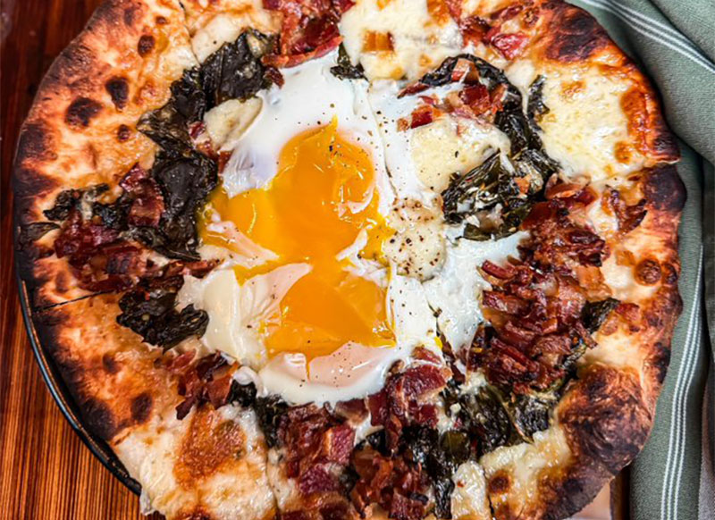 A top-down view of a gourmet wood-fired pizza from Paladar 511, topped with melted cheese, savory meats, and two perfectly cooked over-easy eggs in the center.