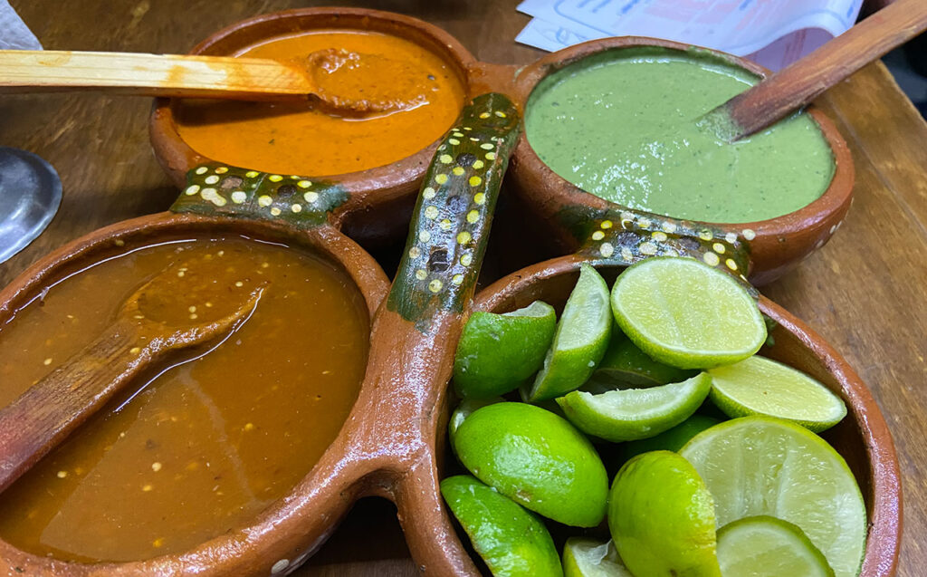 Top-down view of a clay dish filled with three different salsas—green, orange, and dark red—and fresh lime wedges, with wooden spoons resting in the sauces.