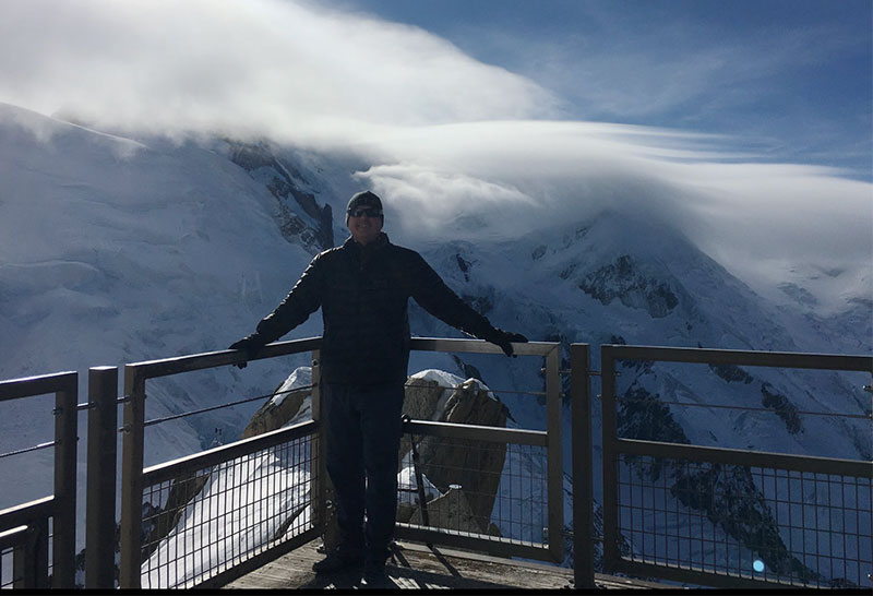 Corey stands on a high-altitude observation deck in Chamonix, France, with his arms outstretched, framed by dramatic white clouds and the towering, snow-covered peaks of the Mont Blanc massif in the background.