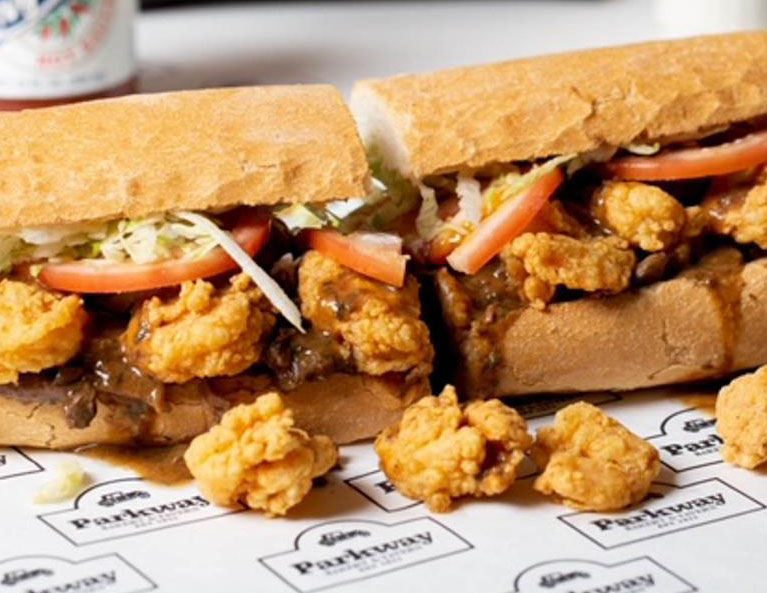 A close-up of a legendary Parkway Bakery & Tavern "surf and turf" po'boy, overflowing with slow-cooked roast beef gravy and golden fried shrimp on crusty French bread.
