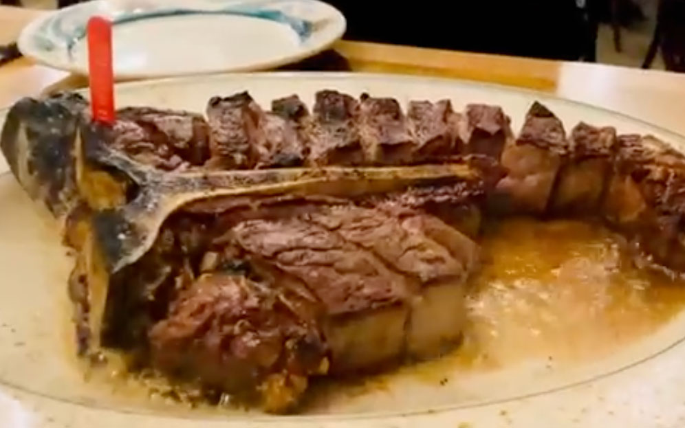A legendary Peter Luger porterhouse steak, dry-aged and sliced, served sizzling on a signature tilted plate with a side of thick-cut bacon.