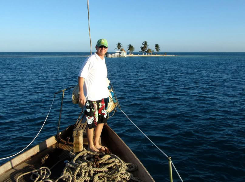 Corey A person stands at the bow of a wooden sailboat, looking out over the vast blue Caribbean Sea under a clear sky during a Ragga Sailing Adventures trip in Belize.