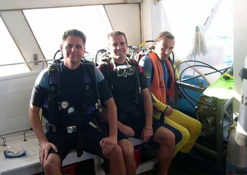Corey, Rob and Ben our drive master in scuba diving gear sitting on a boat and smiling, with the turquoise waters and lush green islands of the Similan Islands in Thailand in the background.