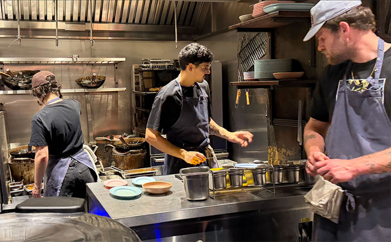 A chef in a black uniform working in an open kitchen at Smoking Goat in London, carefully preparing dishes at a stainless steel counter while customers watch from nearby seats.