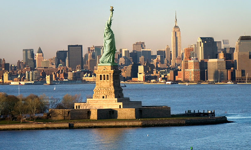 A classic view of the Statue of Liberty standing tall against the Lower Manhattan skyline and the New York Harbor at sunset.
