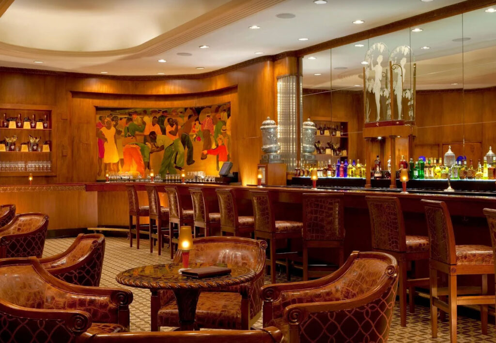 The elegant interior of The Sazerac Bar at the Roosevelt Hotel, featuring its famous Art Deco murals, polished African walnut bar, and plush velvet seating in a dimly lit, historic atmosphere.