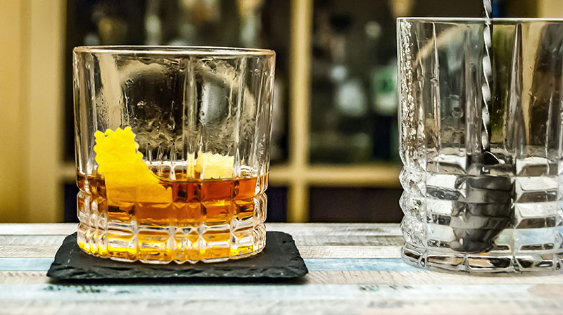 A close-up of a classic Sazerac cocktail in a chilled rocks glass, featuring an amber-colored liquid and a lemon peel garnish, sitting on a dark coaster in a dimly lit, sophisticated bar.