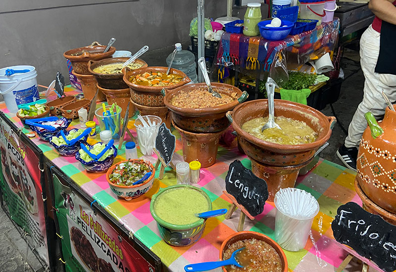 A row of traditional Mexican clay pots (cazuelas) filled with various colorful stews and toppings at a street food stall.