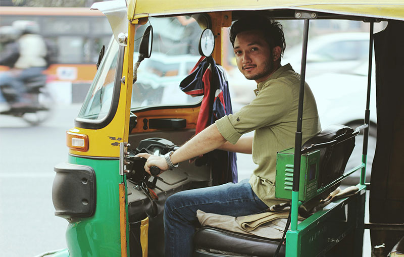 A side-view of a friendly Indian tuk-tuk driver sitting in the front seat of his yellow and green auto-rickshaw in New Delhi, looking back with a warm smile.