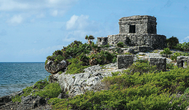 Ancient Mayan stone ruins of Tulum perched on a rocky cliff overlooking a white sand beach and the turquoise Caribbean Sea.