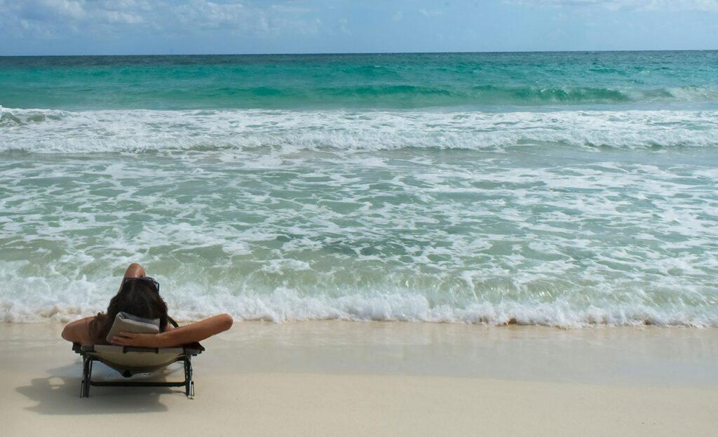 A tranquil view of small waves lapping against the shore at a Tulum beach, with a rustic wooden lounge chair positioned near the water's edge and the vast turquoise Caribbean Sea extending to the horizon.