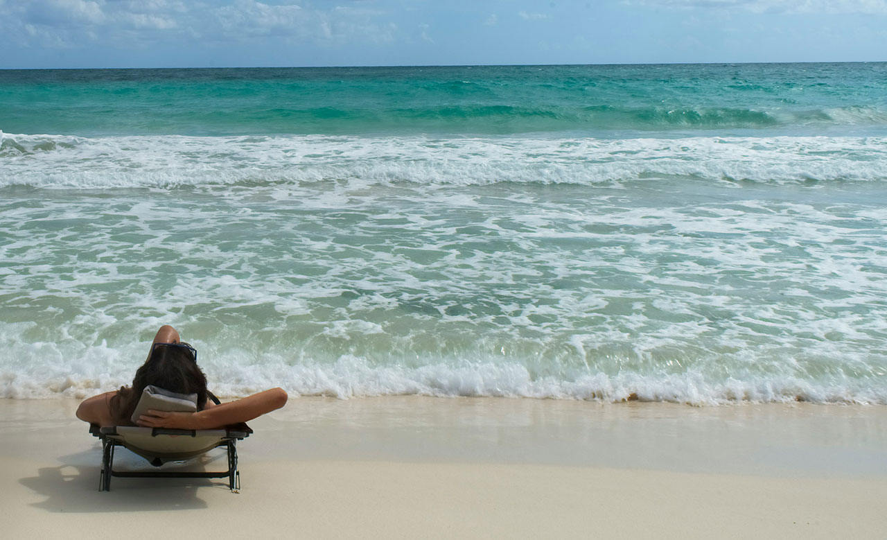 A tranquil view of small waves lapping against the shore at a Tulum beach, with a rustic wooden lounge chair positioned near the water's edge and the vast turquoise Caribbean Sea extending to the horizon.