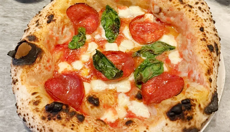 A close-up of a wood-fired Margherita pizza from Una Pizza Napoletana, featuring a charred, blistered crust, melted buffalo mozzarella, fresh basil leaves, and a rich tomato sauce.