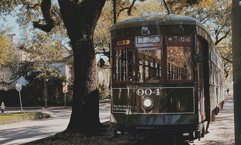 A vintage dark green Perley Thomas streetcar labeled "St. Charles" traveling through a sun-dappled, tree-lined street in the Garden District of New Orleans.