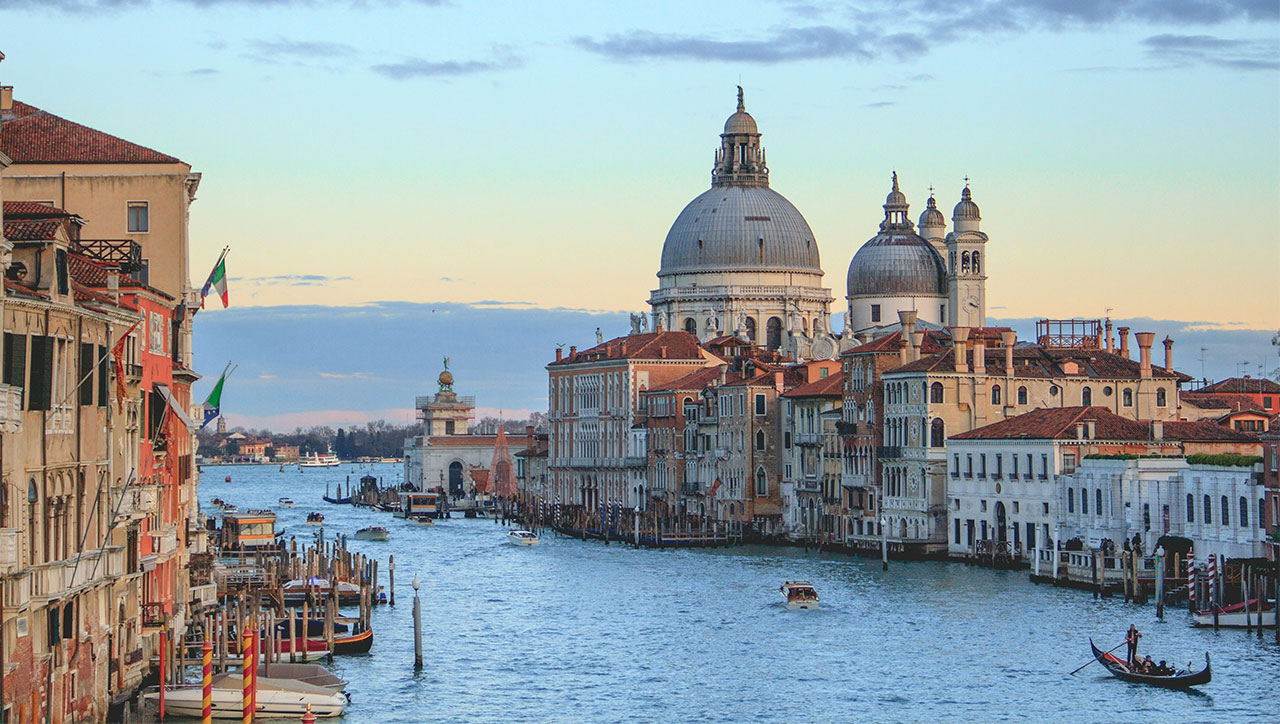 A classic sunset view of the Grand Canal in Venice, looking toward the Basilica di Santa Maria della Salute with historic buildings lining the water and gondolas in the distance.