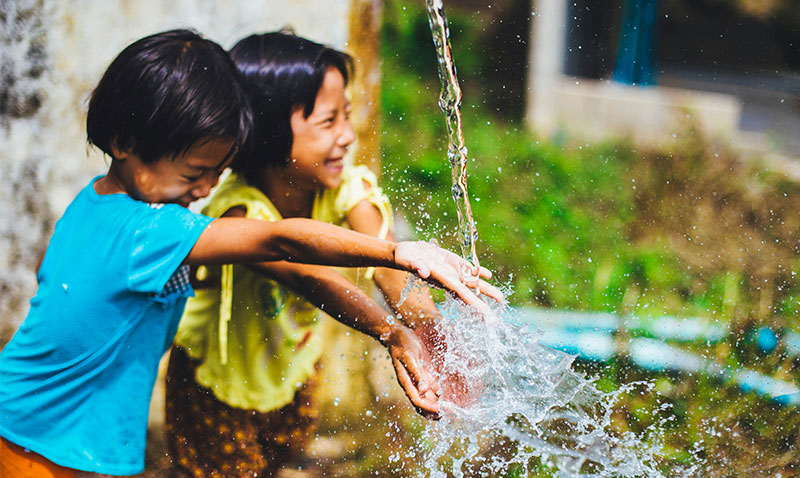 Two young boys in a rural village in Chiang Mai, Thailand, play together with water under a running tap, surrounded by lush green tropical foliage and traditional wooden structures.