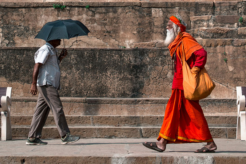 A person holding a black umbrella and a monk in a vibrant orange robe walk along a historic stone path lined with traditional wooden buildings and steps.