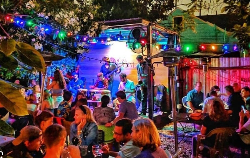 A lush, green outdoor courtyard at Bacchanal Wine in the Bywater, featuring mismatched wooden tables, folding chairs, and string lights overhead, where patrons are enjoying wine and live music in a relaxed atmosphere.