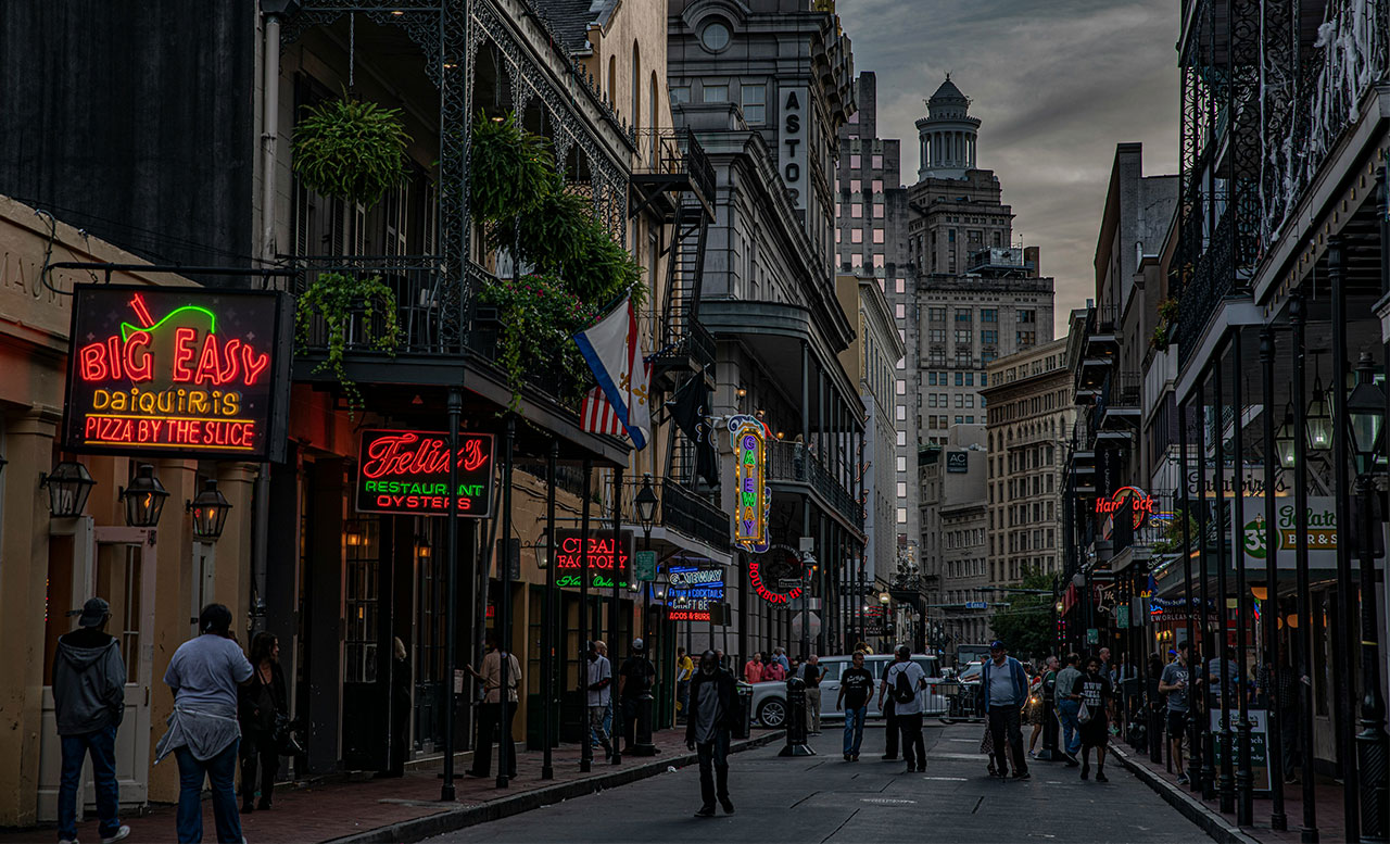 A bustling evening scene on a narrow New Orleans French Quarter street, featuring historic multi-story buildings with wrought-iron balconies, neon signs for bars and clubs, and people walking along the sidewalks.