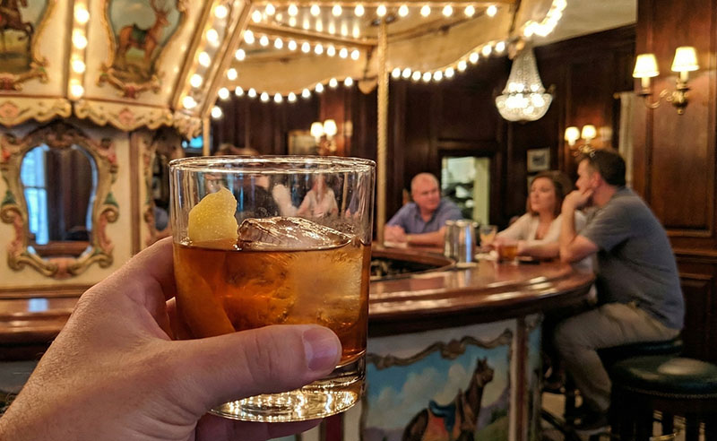A first-person view of a Vieux Carré cocktail being held in front of the ornate, rotating Carousel Bar at the Hotel Monteleone in New Orleans.