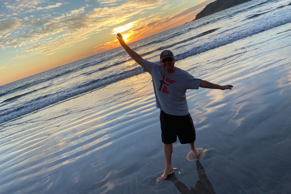 A man in a grey t-shirt, black shorts, and a baseball cap stands on a wet sandy beach at sunset, holding his arms out wide with one hand appearing to "touch" the sun.