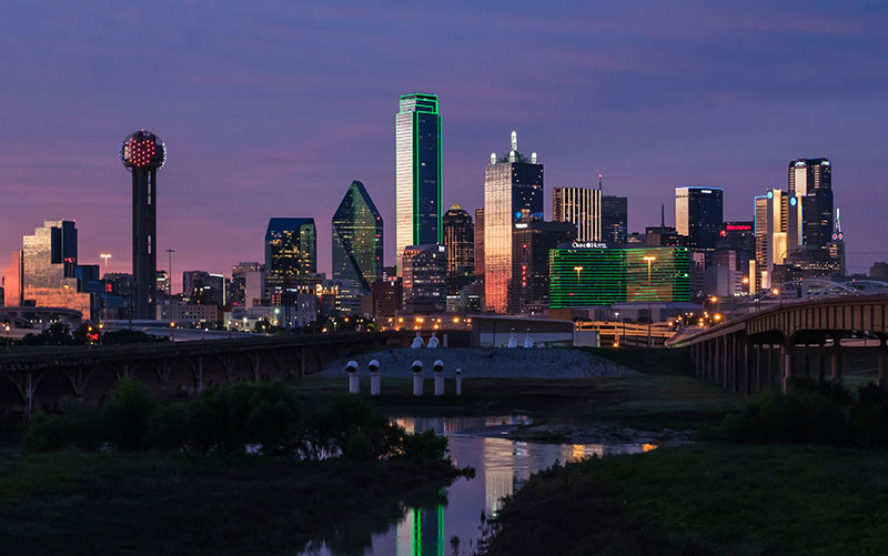 A night view of the Dallas, Texas skyline featuring the illuminated Reunion Tower and colorful skyscrapers reflecting in the water.