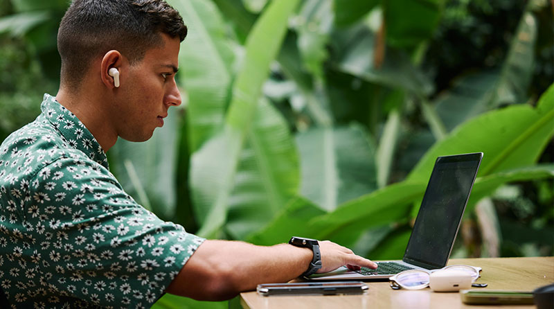 A side profile of a young man working on a laptop in a lush, outdoor tropical setting, wearing a green patterned shirt and headphones.