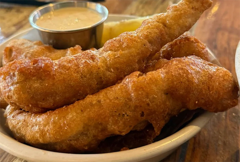 A close-up of a platter featuring golden-brown fried fish fillets served with a small side of dipping sauce at Pêche Seafood Grill.