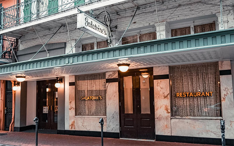 he iconic exterior of Galatoire’s Restaurant on Bourbon Street, featuring its historic green-and-white striped awnings, classic neon signage, and large arched windows reflecting the vibrant energy of the French Quarter.