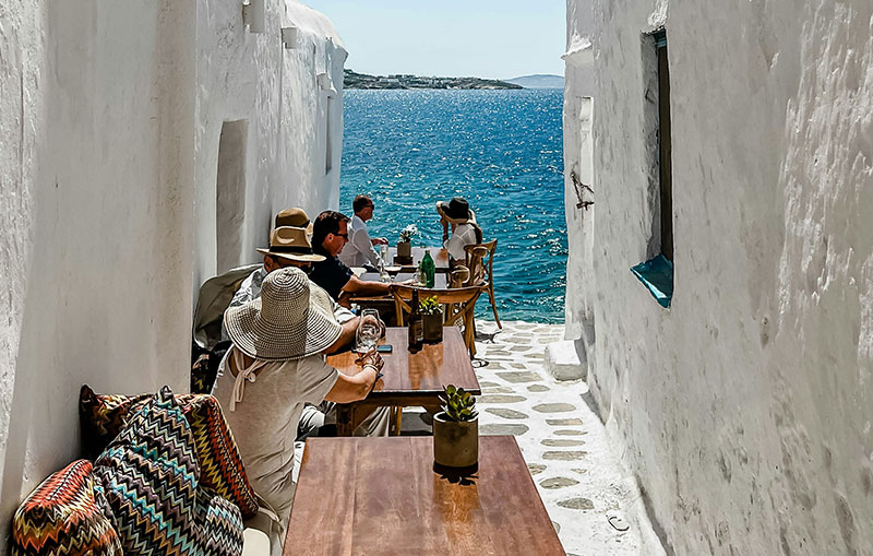 sun-drenched outdoor cafe in a narrow white-walled Greek alleyway, with patrons sitting at wooden tables overlooking a calm blue sea.