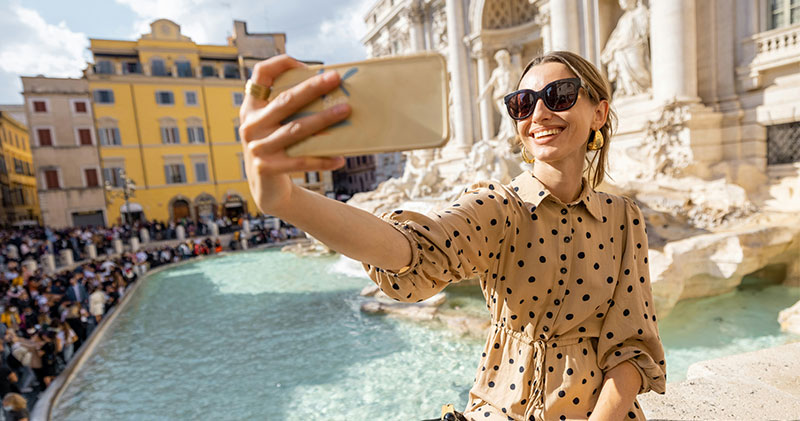 A smiling woman with sunglasses and a polka-dot dress holds up a smartphone to take a selfie in front of the crowded Trevi Fountain in Rome, Italy